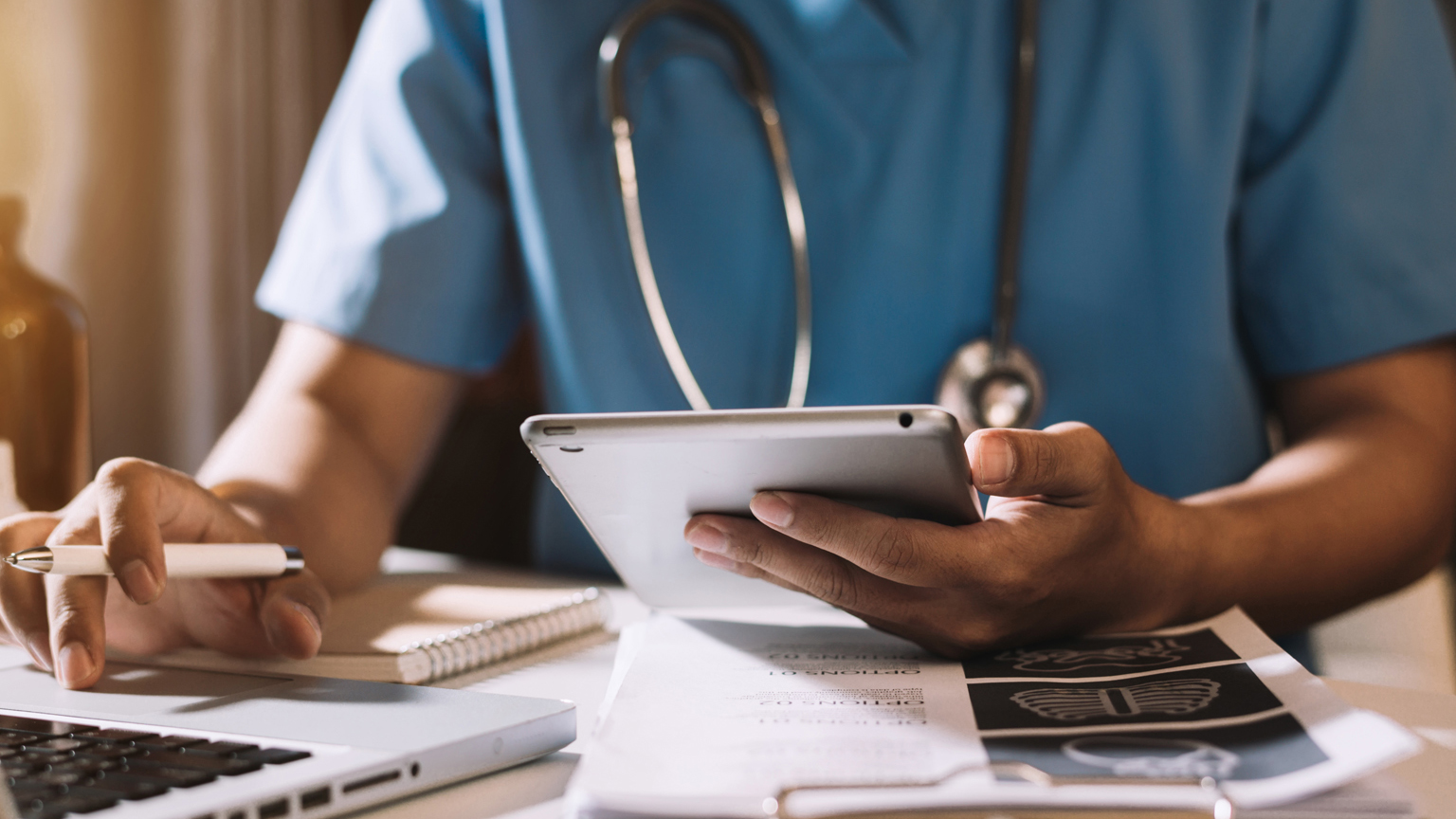 Healthcare professional working at desk on laptop and clipboard of papers with tablet and pen in each hand