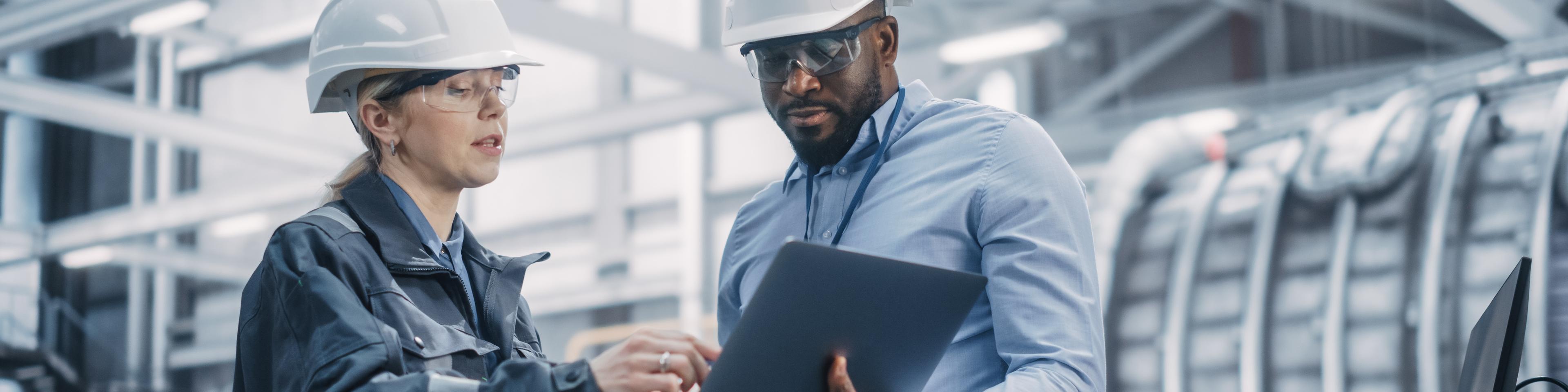 Two Diverse Professional Heavy Industry Engineers Wearing Safety Uniform and Hard Hats Working on Laptop Computer. African American Technician and Female Worker Talking on a Meeting in a Factory.