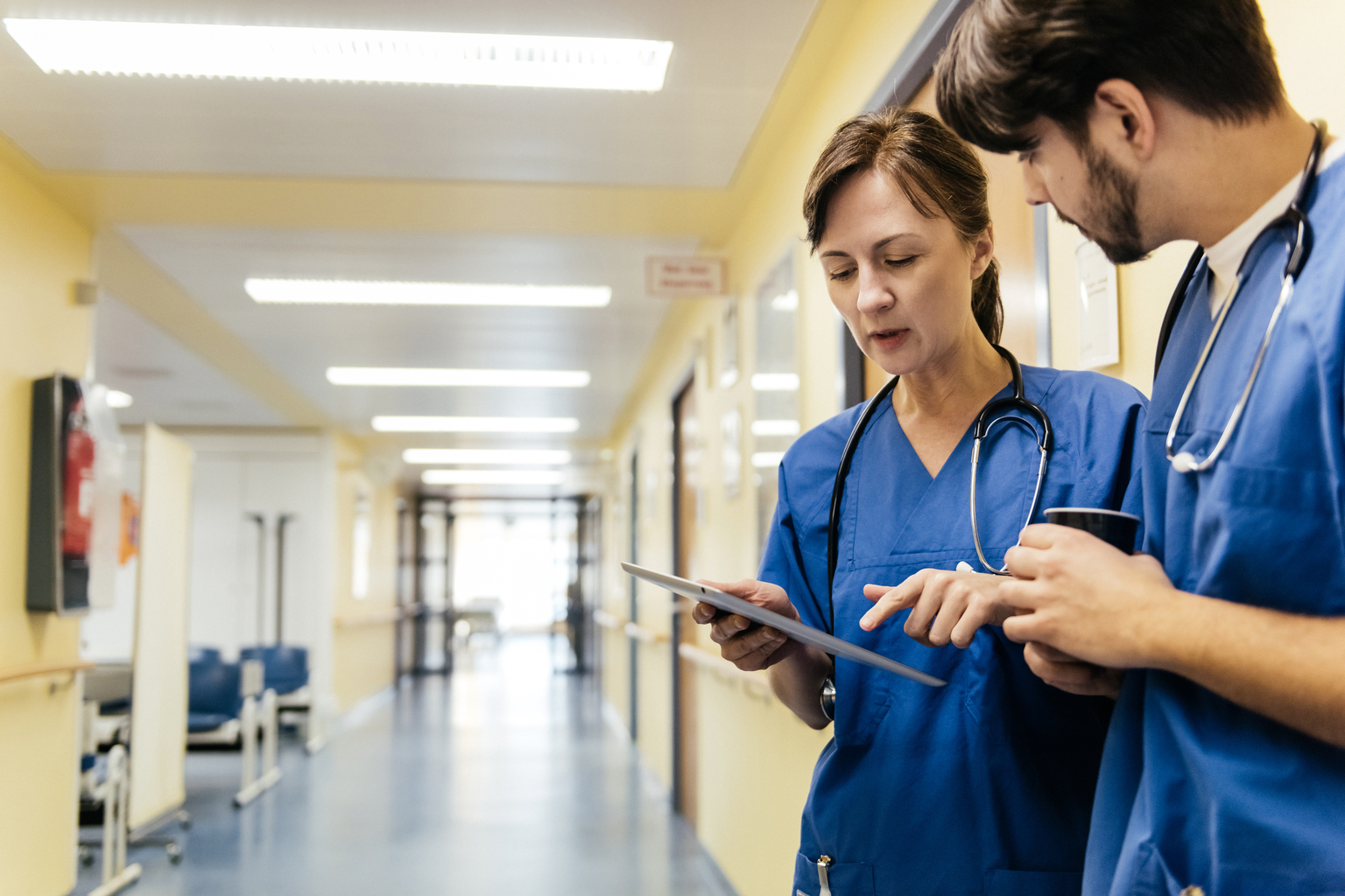 Mature white female doctor and her younger male colleague discussing patient record on a digital tablet while standing in a hospital corridor