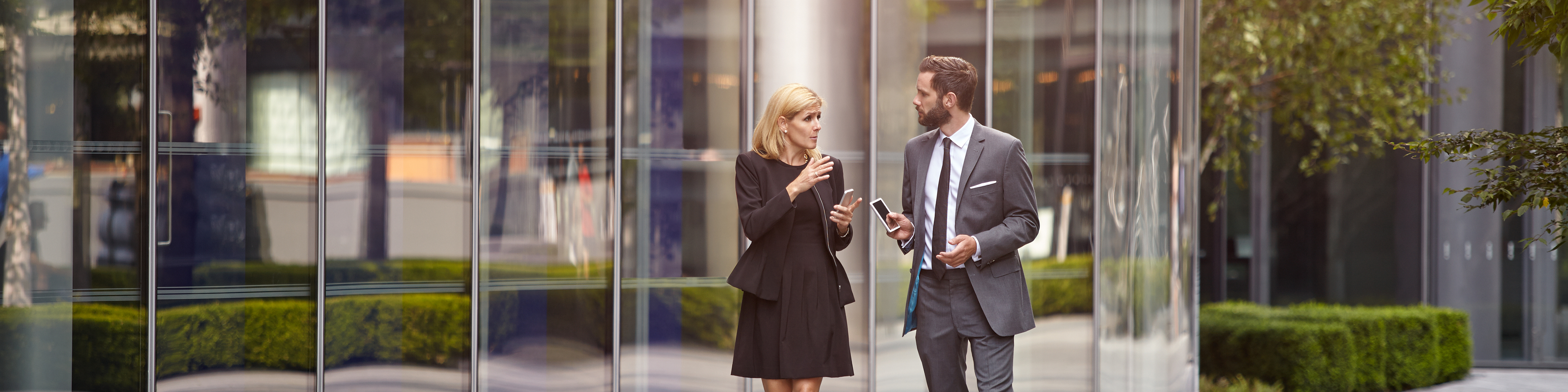 A senior female executive and a coworker outside an office walking and talking discussing plans in a digital tablet