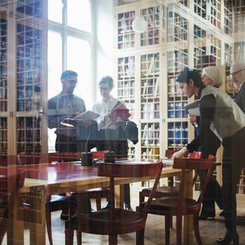 Five people in formalwear including active seniors to discuss a case near the bookshelf at law firm in Europe