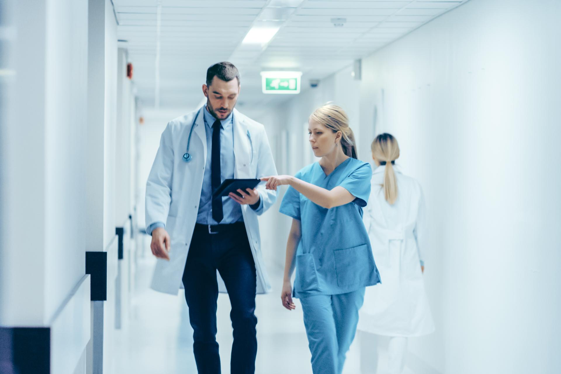 Female surgeon and male doctor walk through hospital hallway, consulting digital tablet computer while talking about patient's health