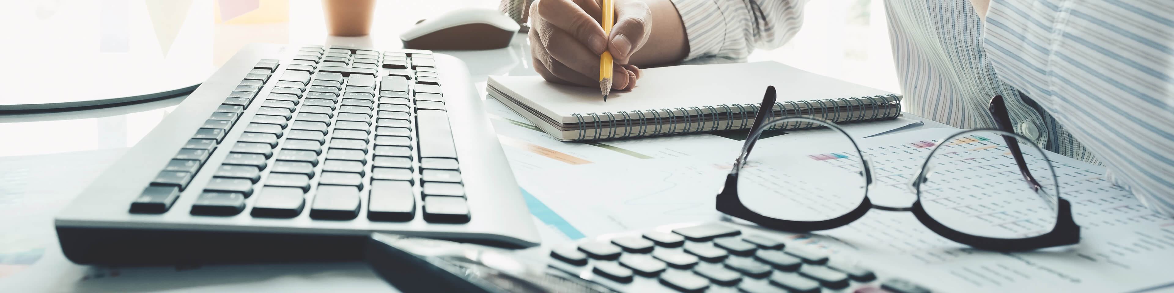 desk with black and white keyboard, glasses, calculator, and paper sitting on top. Man's hand writing on a pad of paper with a yellow pencil.