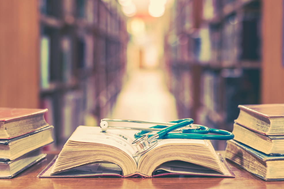 Old book in library with stethoscope on open textbook, stack piles of texts on reading desk, and aisle of bookshelves in study class room background