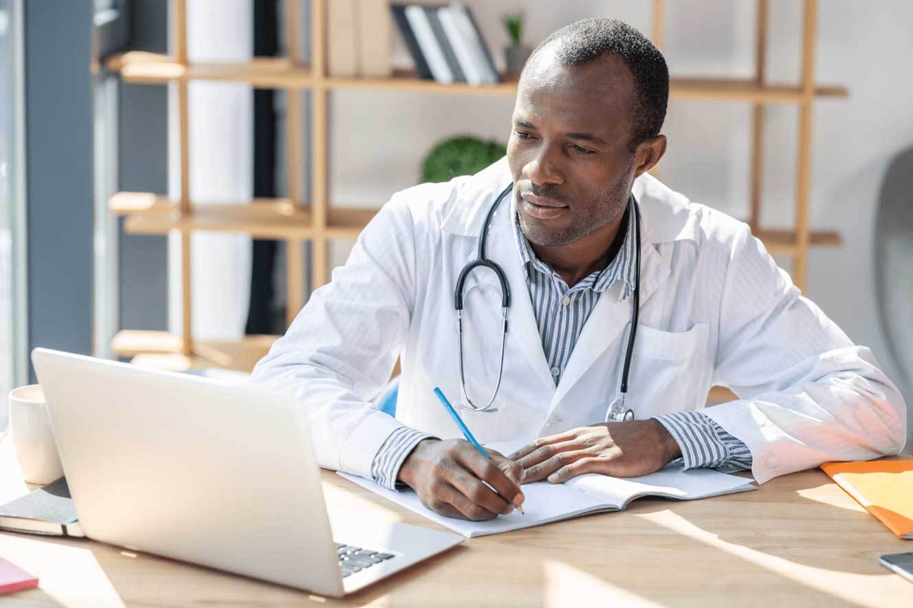 Black doctor looking at computer and taking notes while sitting at workplace