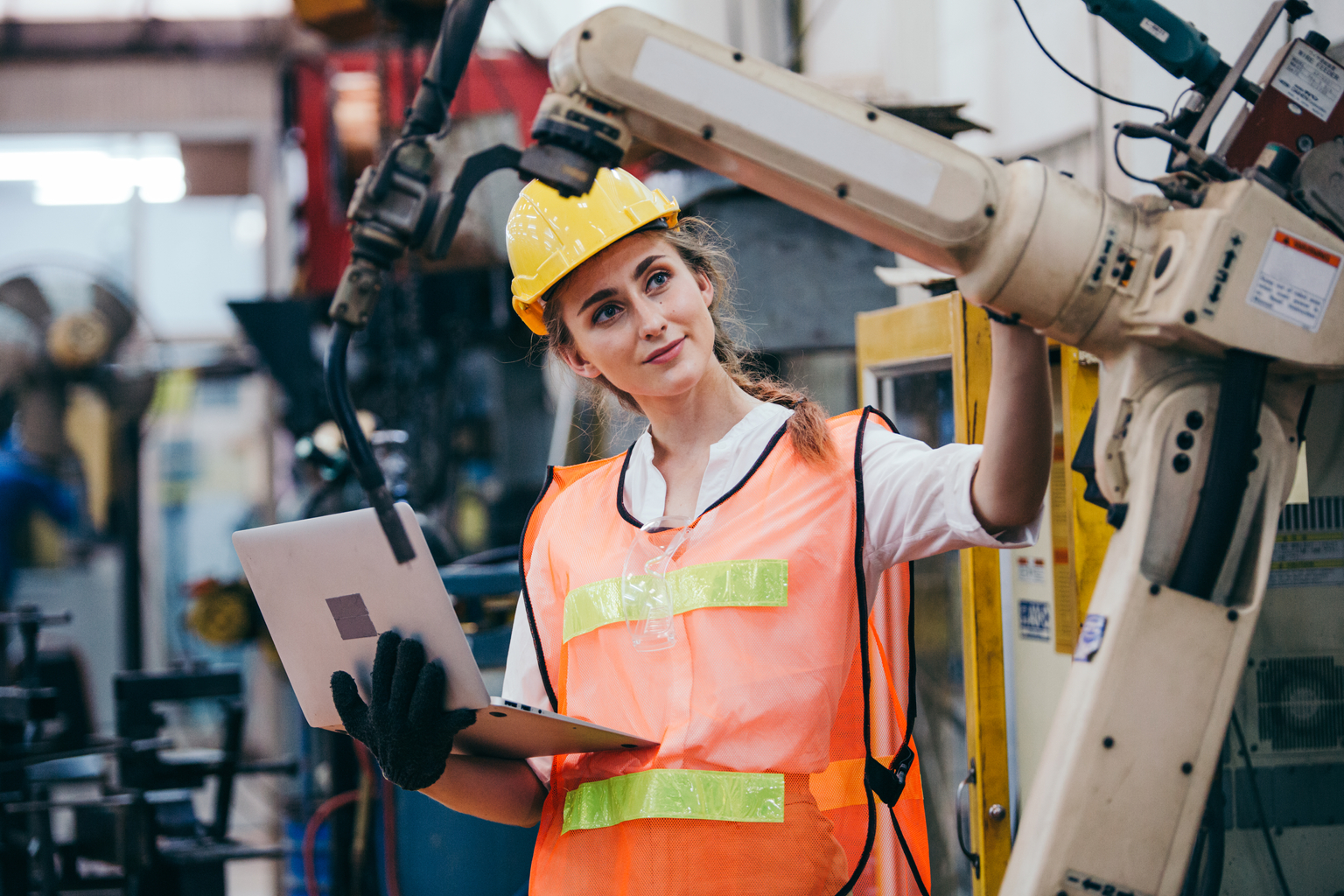 Female industrial engineer or technician worker in hard helmet and uniform using laptop checking on robotic arm machine. woman work hard in heavy technology invention industry manufacturing factory