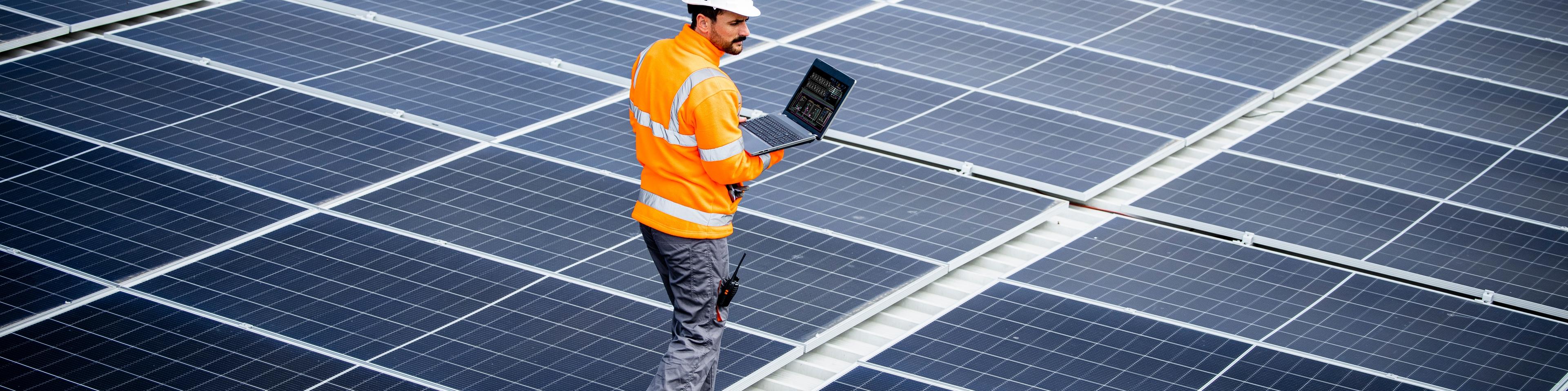 Renewable energy engineer walking through large solar power plant and checking electricity production on his laptop computer. Sustainable energy source and eco friendly technologies.