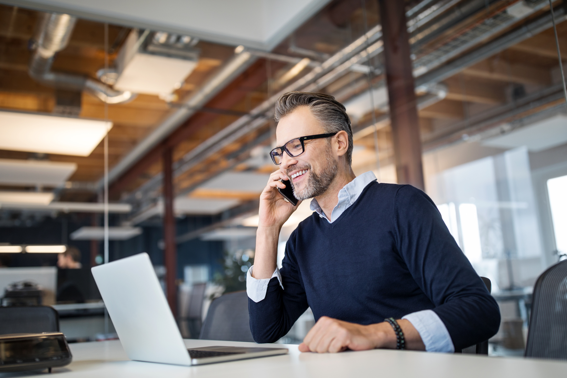 Man with black glasses, blue collared shirt, black bead bracelet and navy sweatshirt sitting down and smiling on the phone, Q3 2021, TAA NA US - Preparer