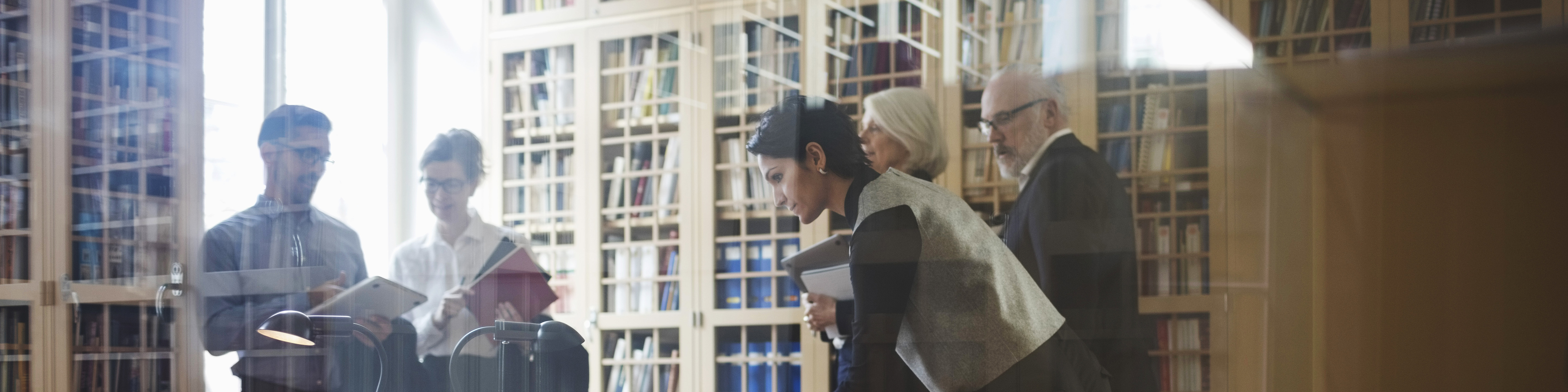 Five people in formalwear including active seniors to discuss a case near the bookshelf at law firm in Europe