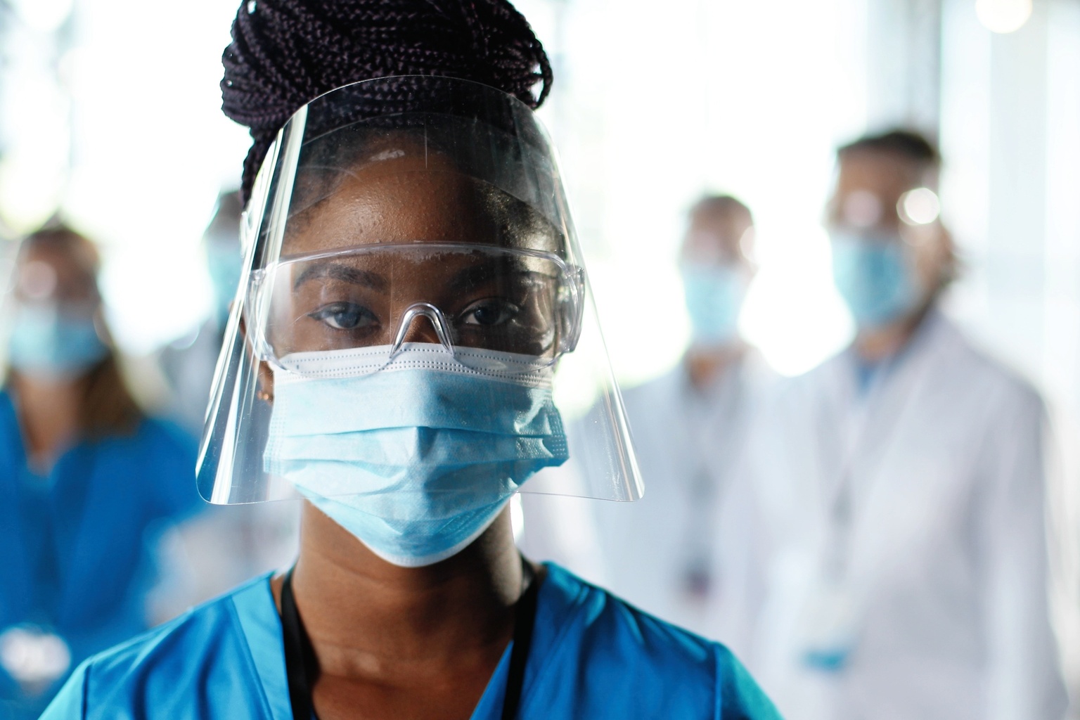 Close up of African American woman doctor or nurse in medical mask, goggles, and face shield in hospital looking to camera, additional doctors in blurred background