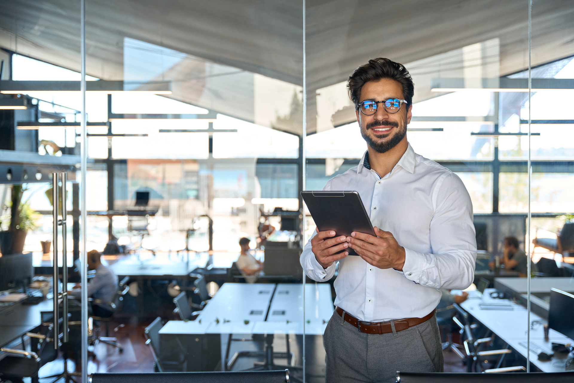 Smiling young business man entrepreneur using tablet standing in office at work. Happy male professional executive ceo manager holding tab computer looking away and thinking of tech data.