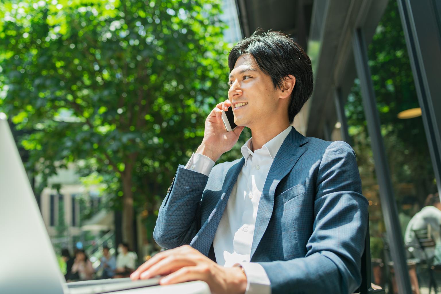 A young Asian businessman on a smartphone outside, his laptop in front of him