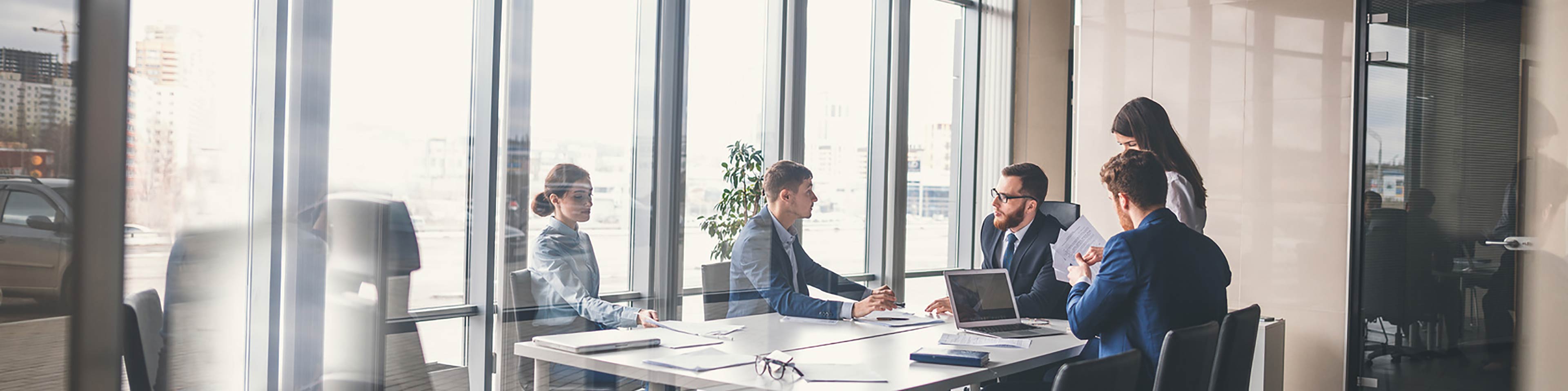 business people sitting around a desk in an office with a glass wall.