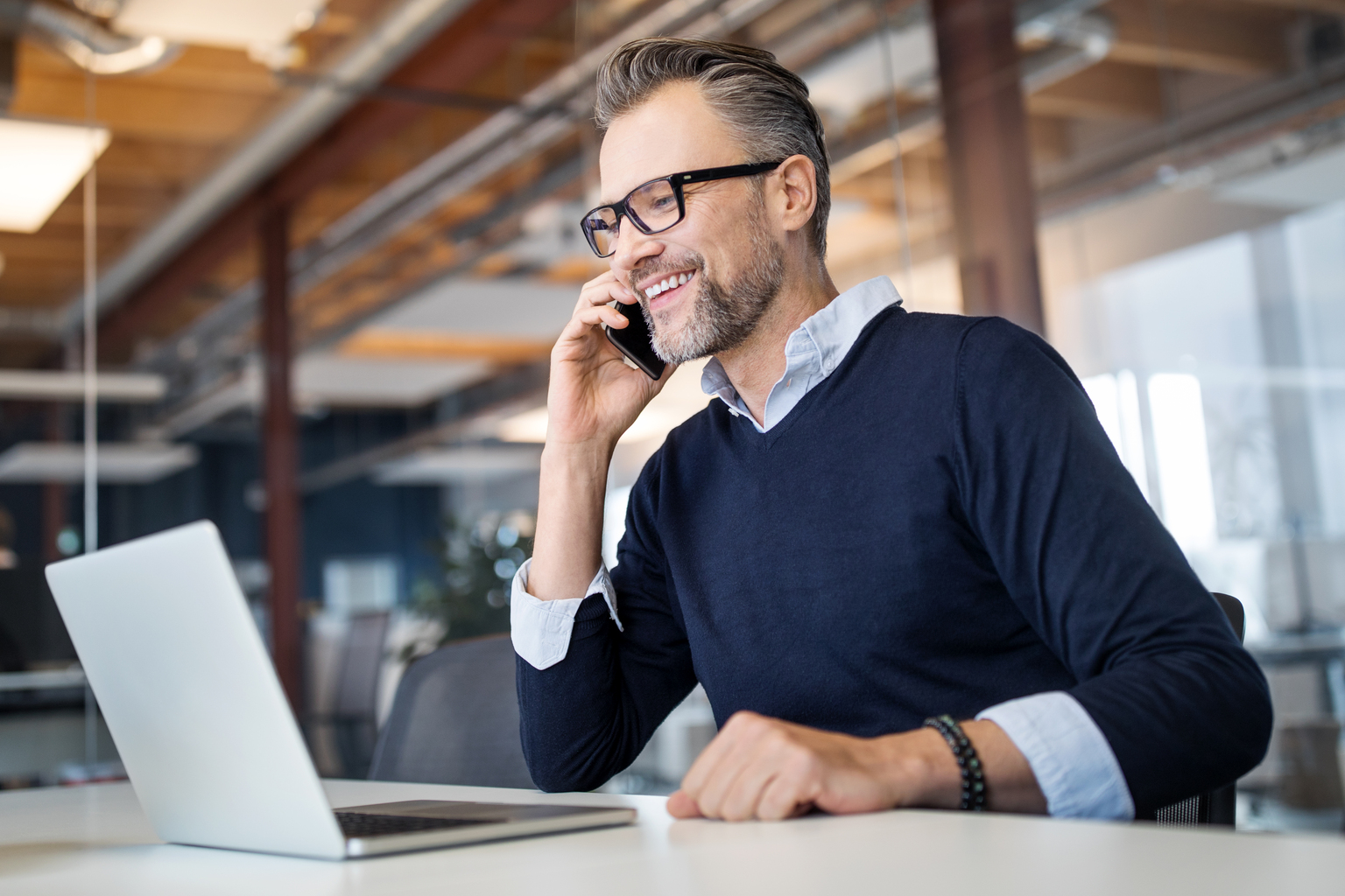 Mid adult business professional sitting at table with laptop and talking on mobile phone. Businessman working in office.