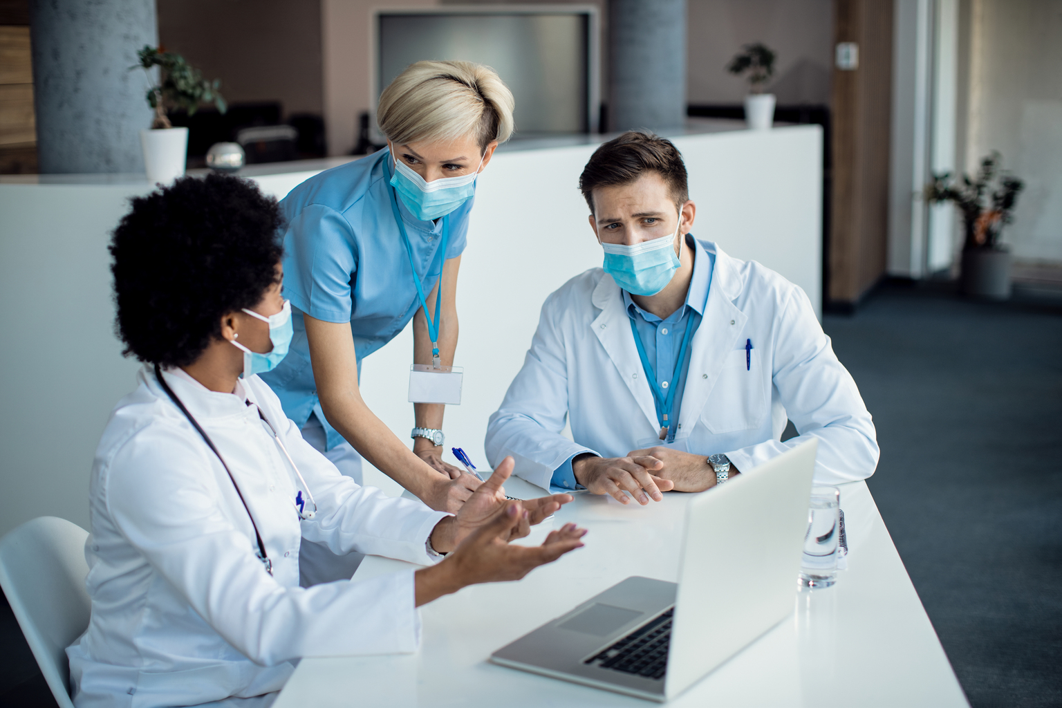 Group of mask wearing medical professionals talking while using laptop during a meeting in hospital