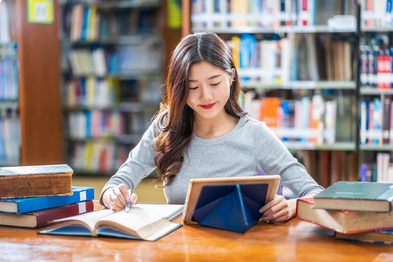 Asian young Student using technology tablet in university library with various books and stationary over the book shelf background