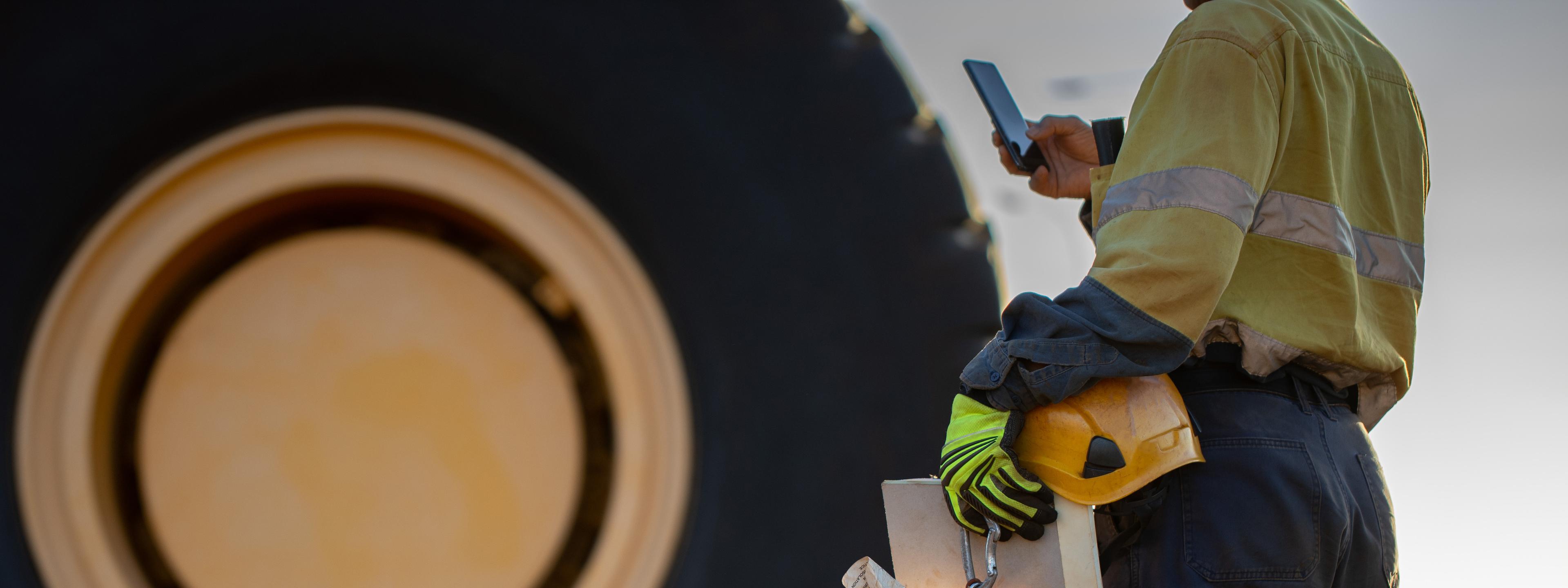 Haul truck inspector wearing work uniform safety glove holding hard hat danger tags personal locks inspection pre operation book inspecting taking pic with cell phone defocused haul truck background