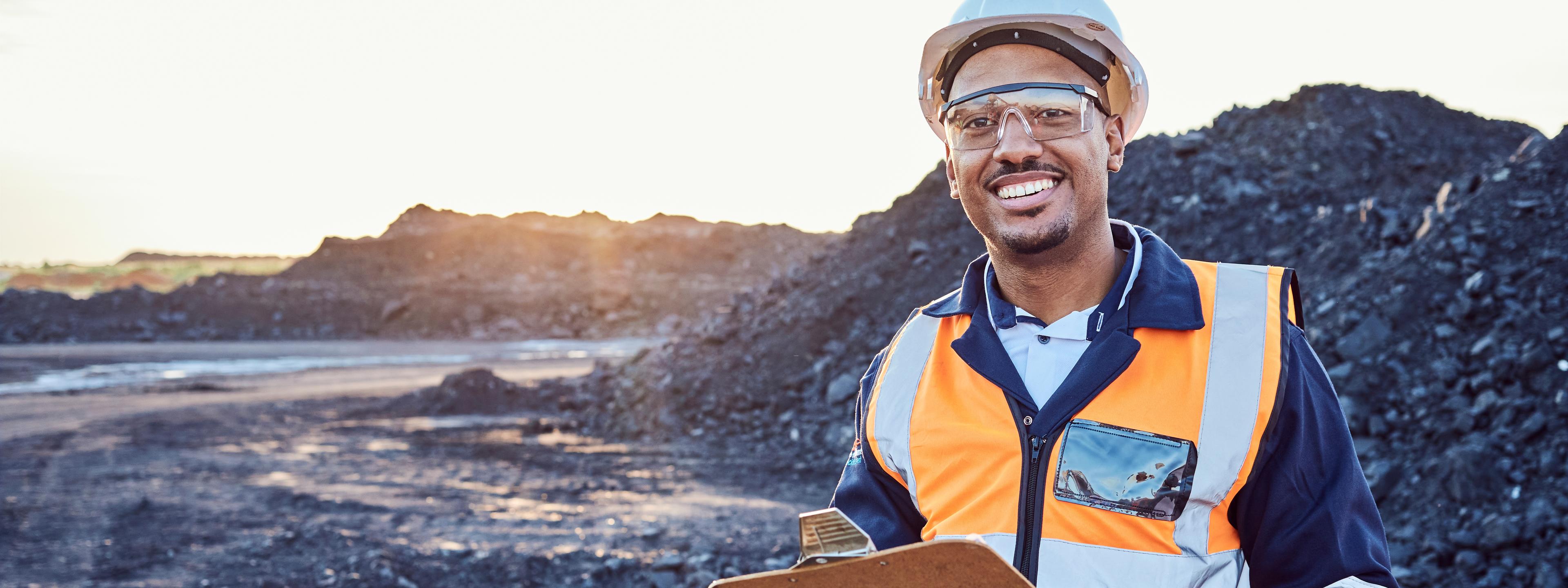 A young African mine worker wearing protective wear is looking at the camera while holding a clipboard with coal mine in the background