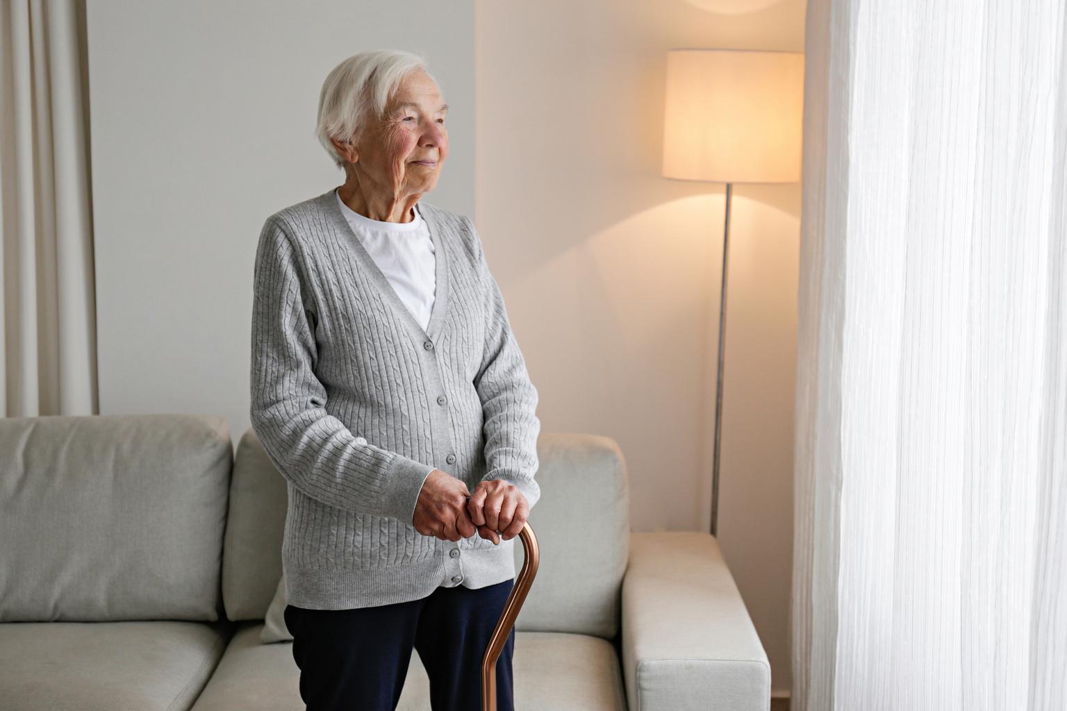 Senior woman standing by the window with walking cane in apartment