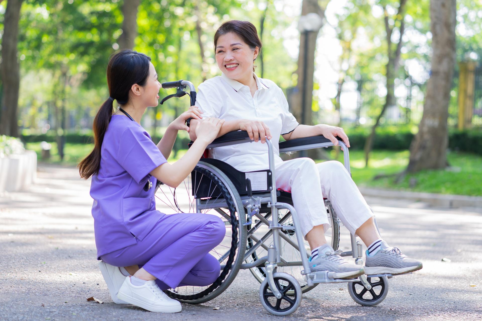 A nurse helps a patient in a wheelchair navigate an outdoor courtyard, both are smiling and having a nice time.