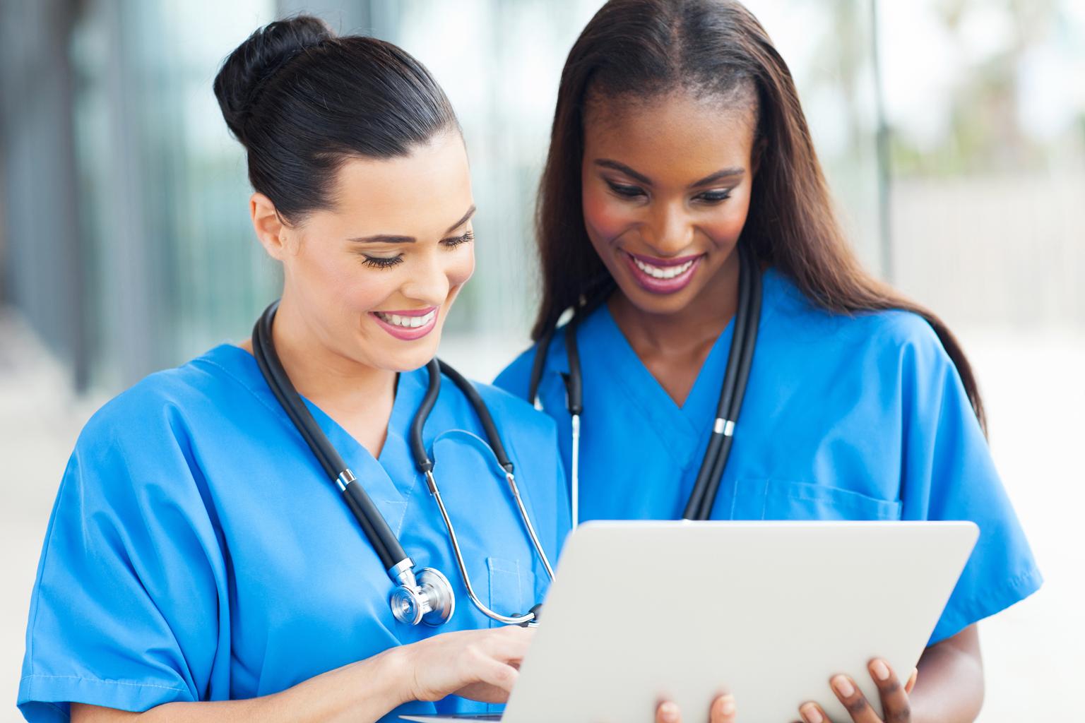 Two nurses in blue scrubs, with stethoscopes around their necks, use tablet to review patient information