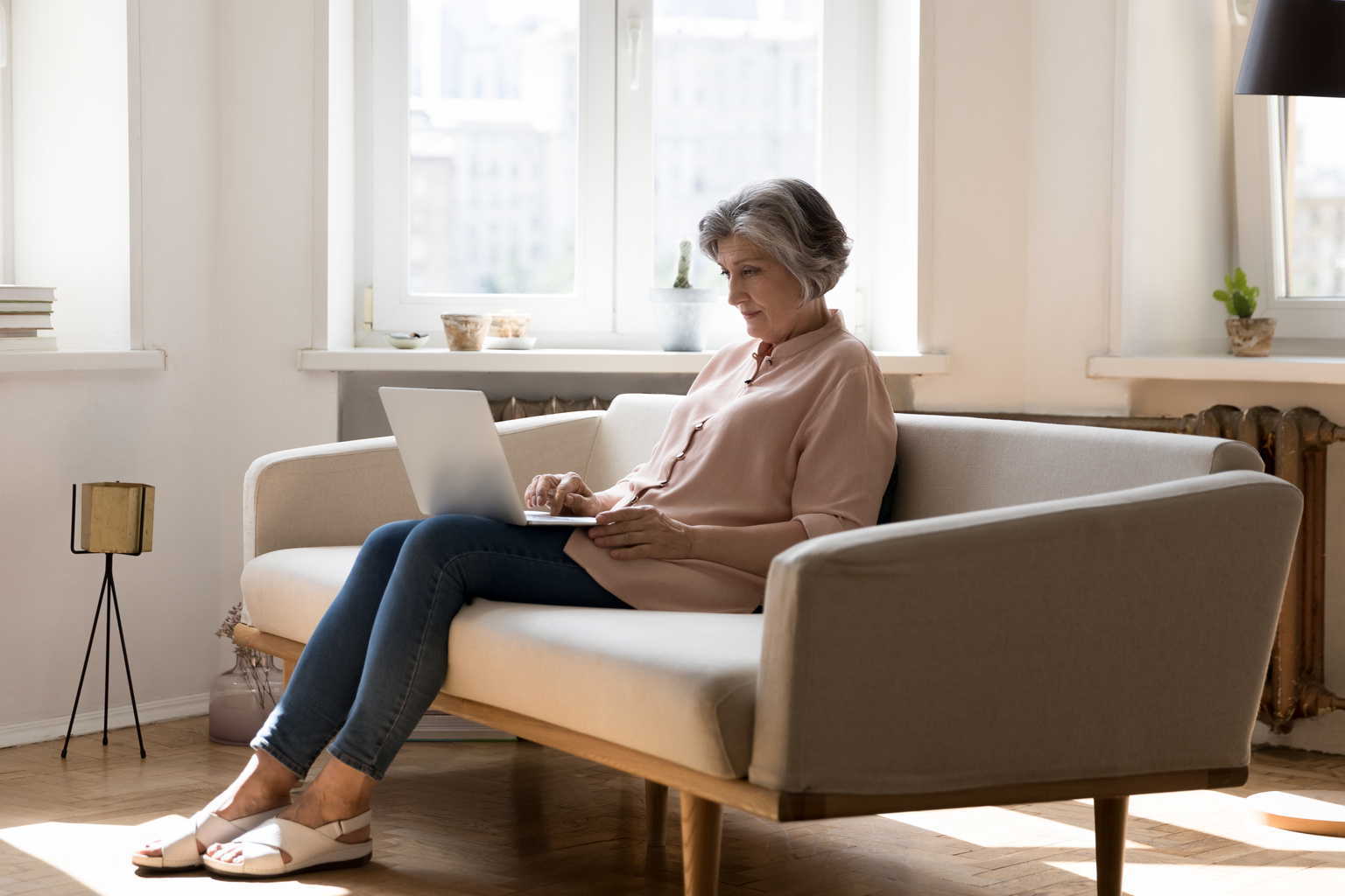 Middle aged grey haired freelance woman using laptop at home, sitting on couch, working on online project