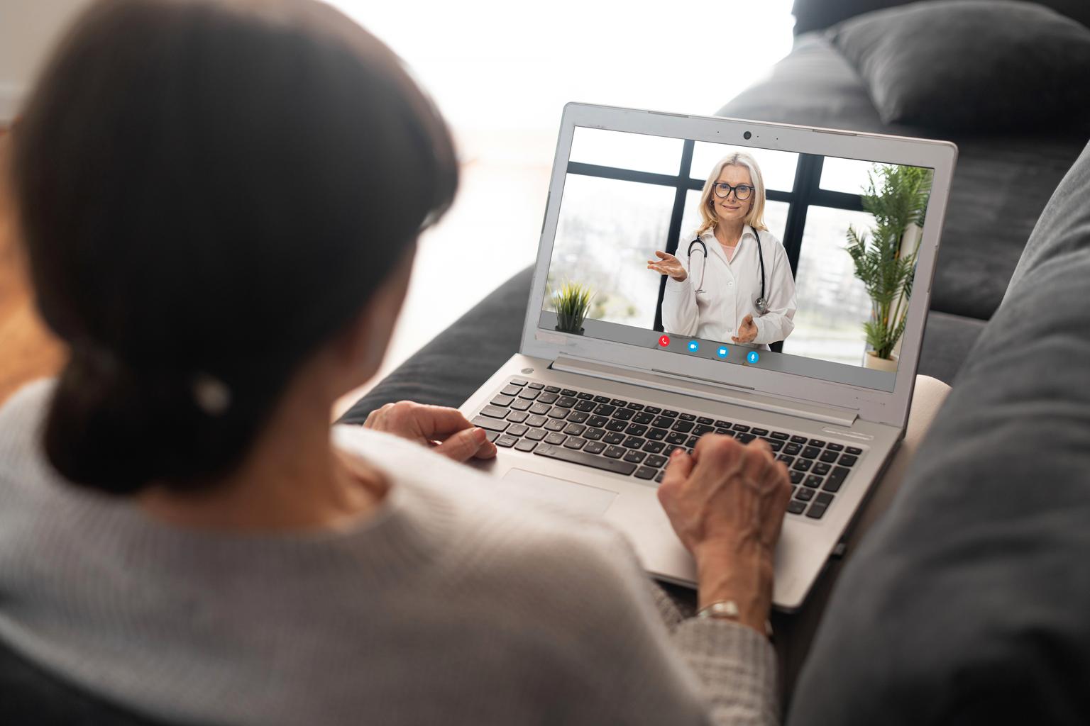 Mature senior lady holding a laptop and talking to a doctor online from home, having a virtual medical check-up with the medical specialist on a webcam