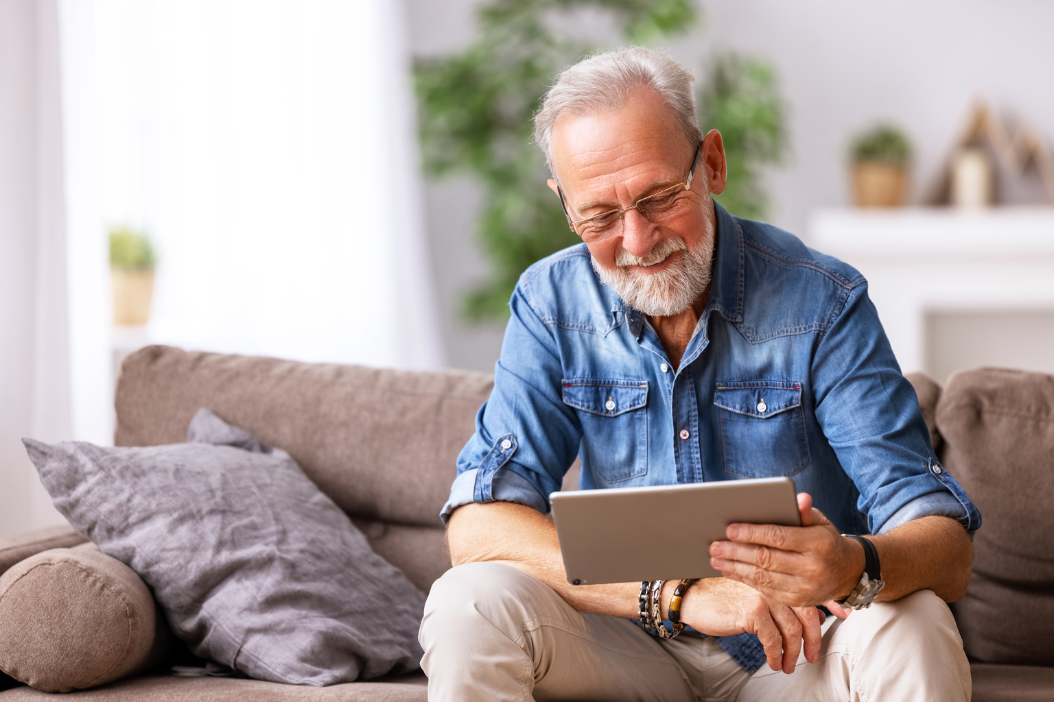 Cheerful aged man using a tablet on a couch.