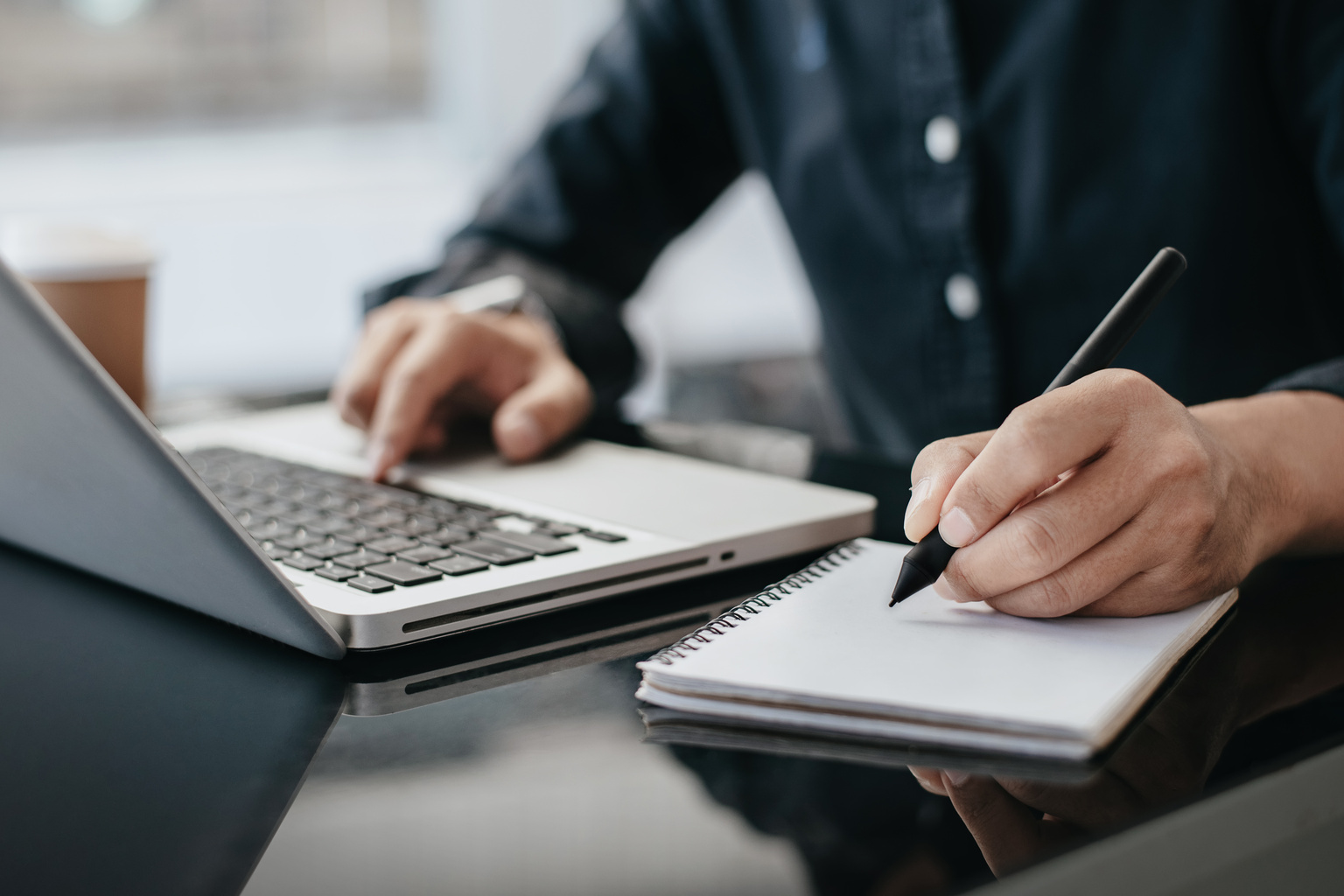 Male businessman using a laptop computer and writing notes in the notebook at the office desk in the morning