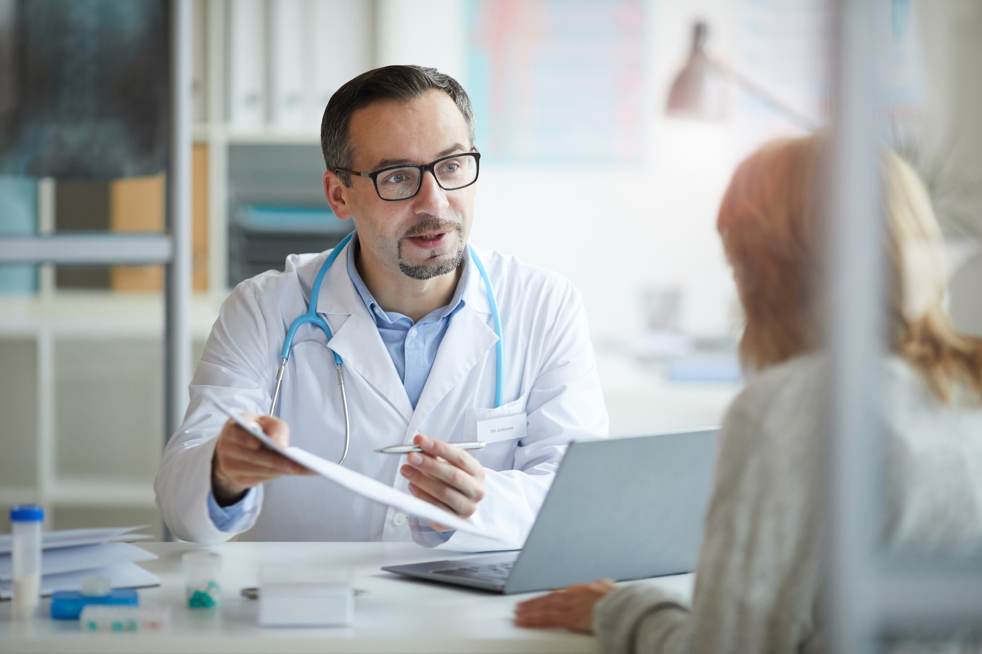 ​​​​​​​Male doctor in a white coat pointing at a document with treatment and showing it to his patient while they are sitting at the table.