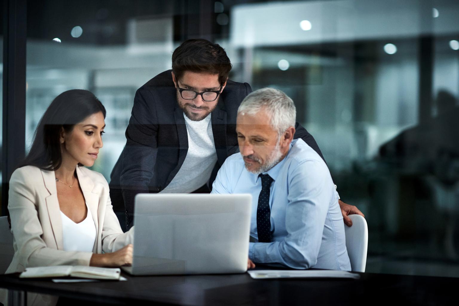 two men and a woman sitting at a table looking at a laptop