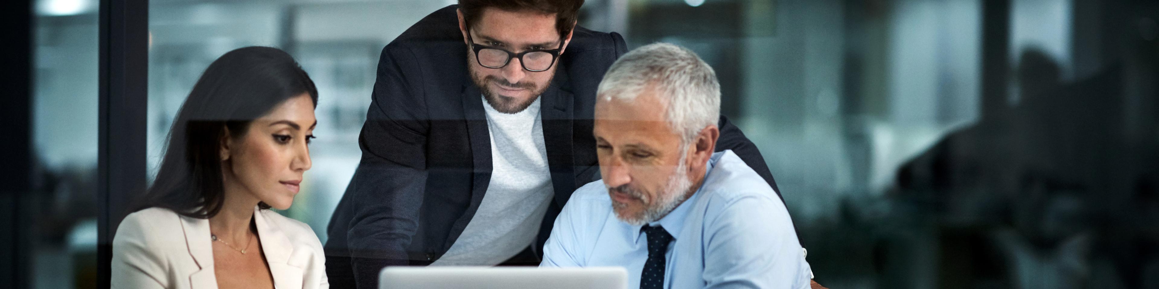 two men and a woman sitting at a table looking at a laptop