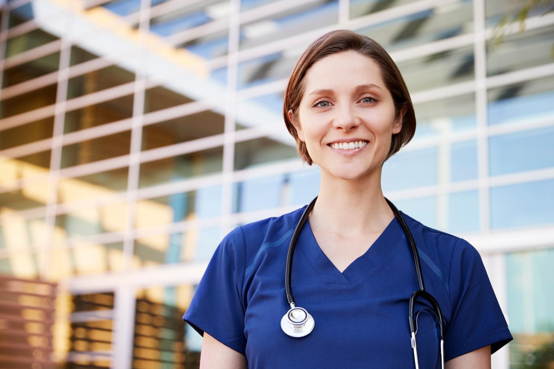 Healthcare worker wearing blue scrubs, smiling at the camera, standing just outside of hospital