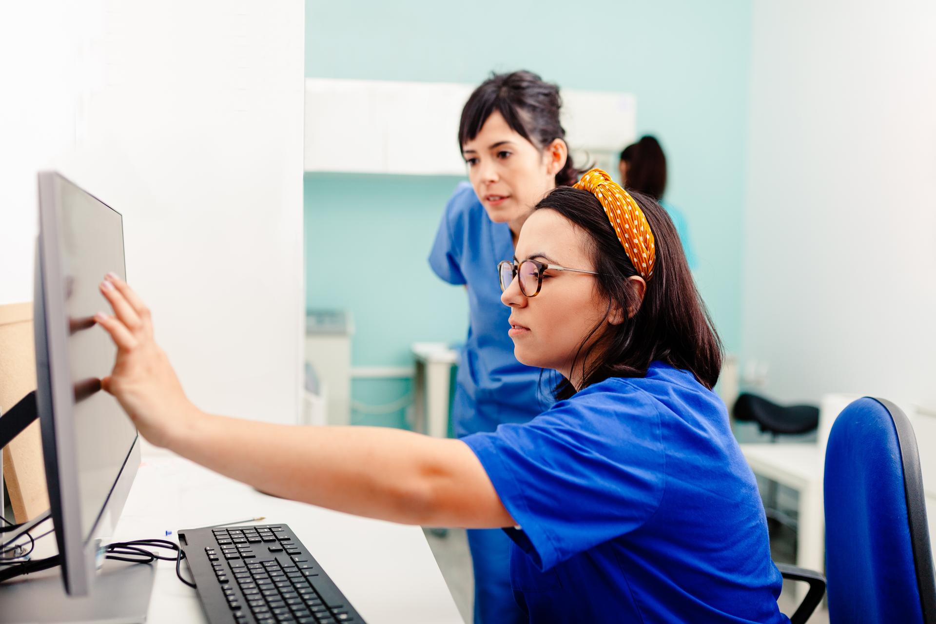 Nurse wearing orange headband and blue scrubs shows colleague how to use EHR