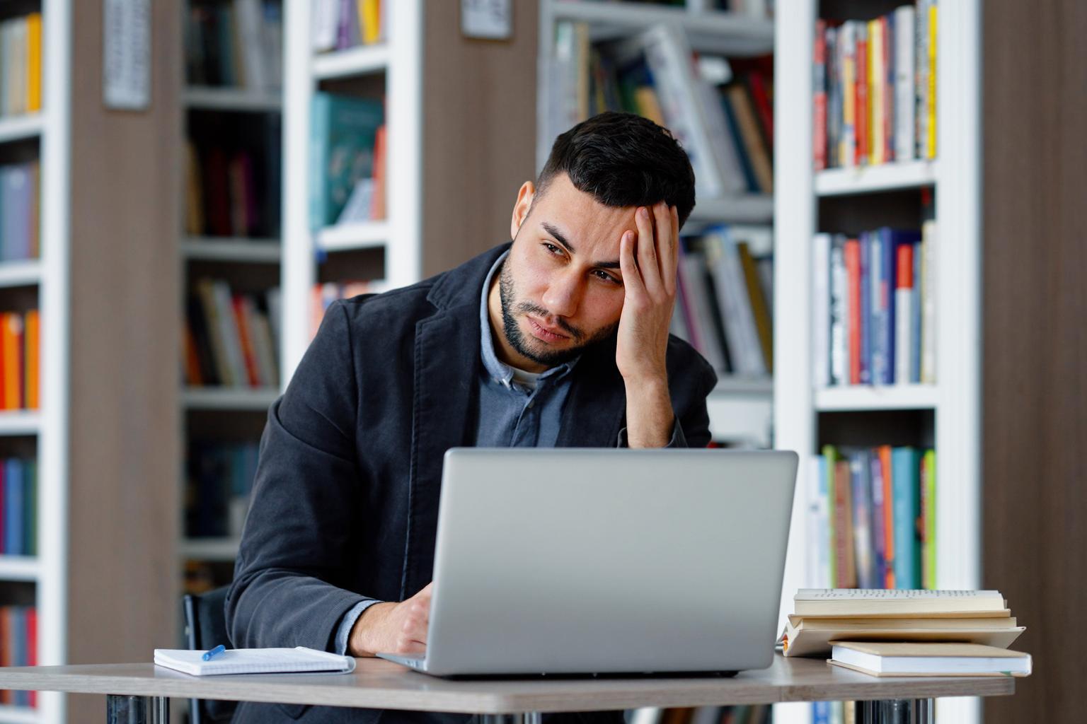 Portrait of man suffering migraine massaging head, working with a laptop in a library