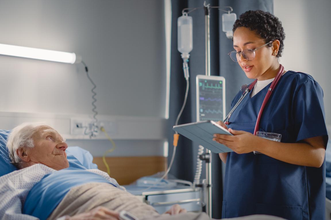 Nurse uses tablet to refer to patient notes as she checks in on elderly patient at the bedside.