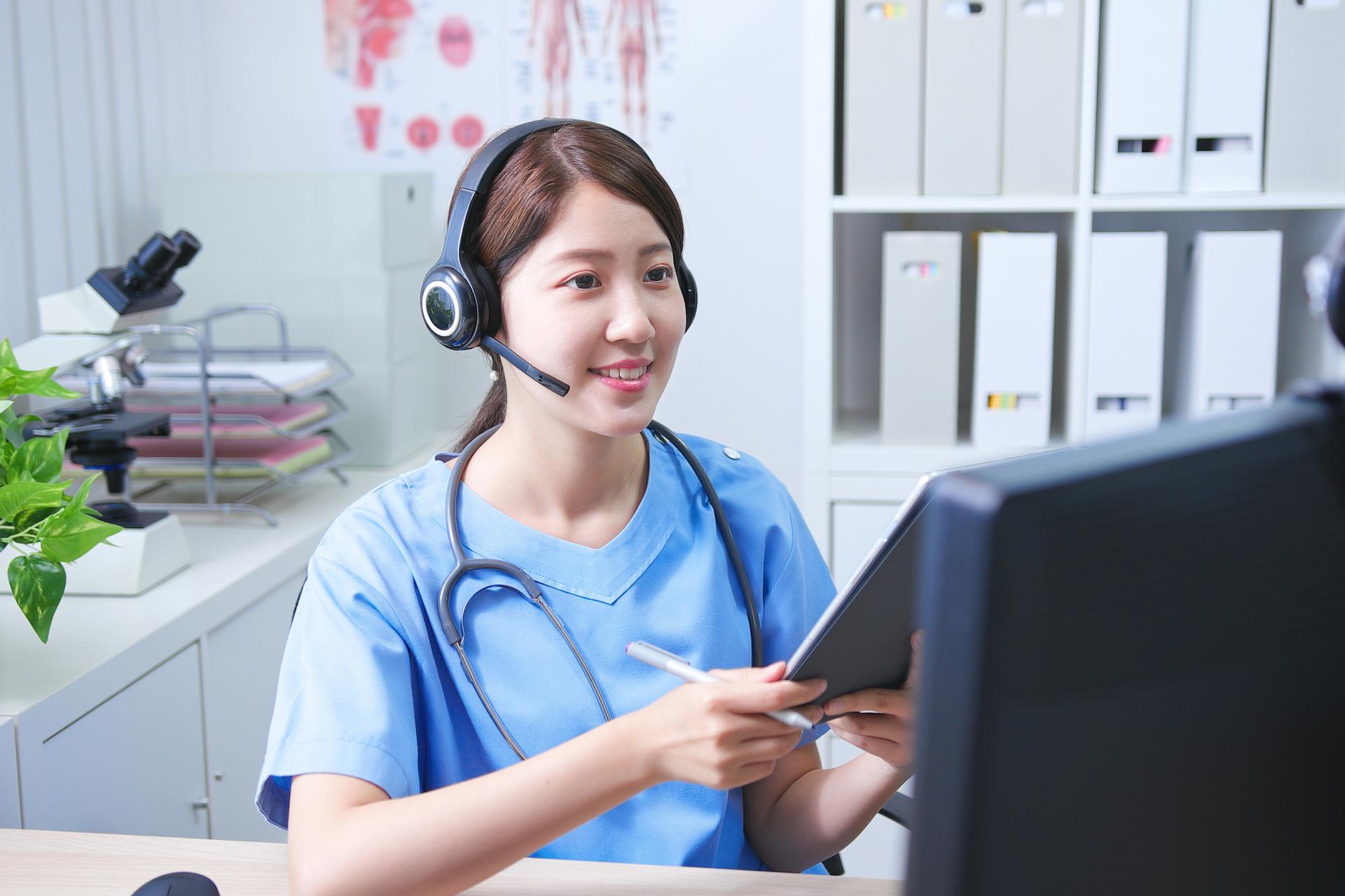 A virtual nurse in scrubs and at her desk, using a computer to provide telehealth healthcare based on virtual care models.