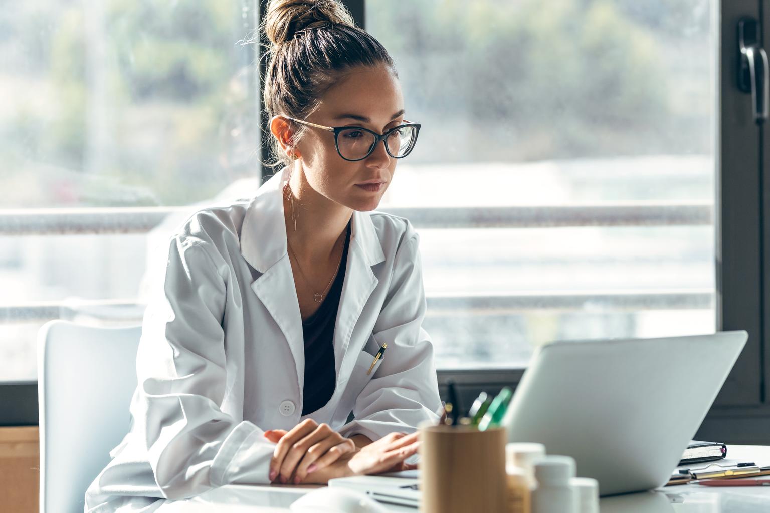 A female doctor explaining medical treatment to a patient through a video call, using a laptop and earphones in a consultation setting