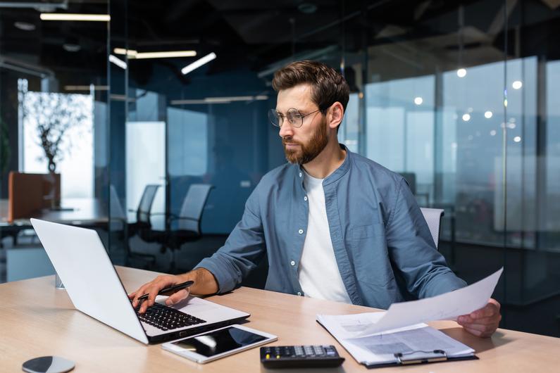 A man sitting in the office, holding documents, checking accounts, and typing on a laptop.