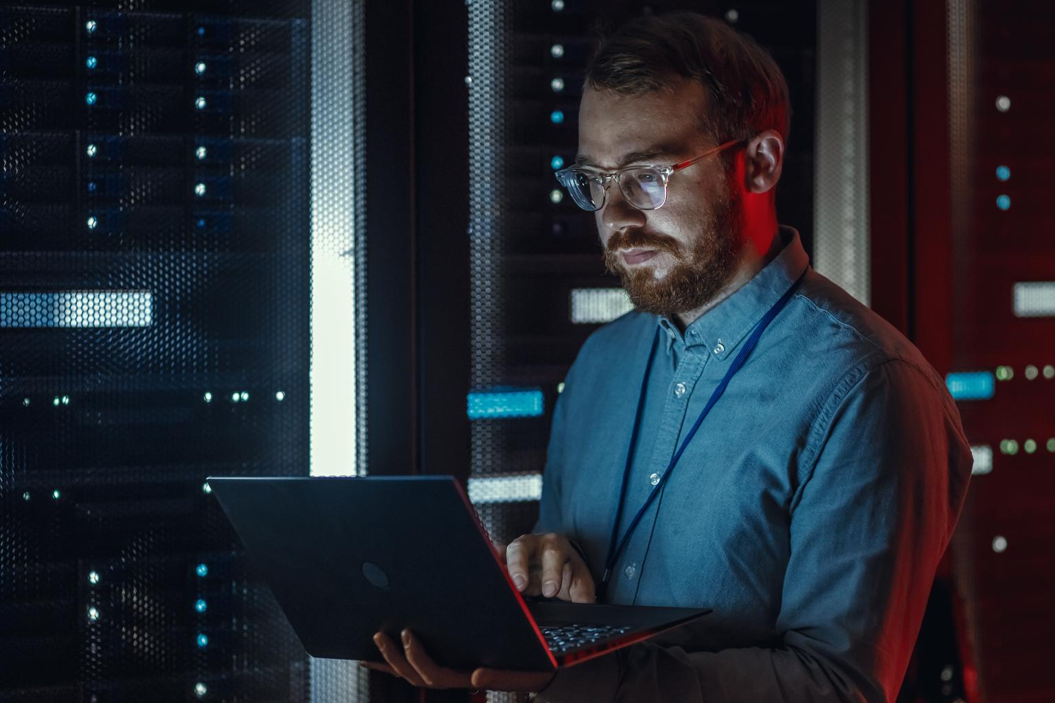 Bearded IT Specialist in Glasses is Working on Laptop in Data Center while Standing Near Server Rack. Running Diagnostics, Doing Maintenance Work. Emergency Red Light from Side Illuminating Specialist