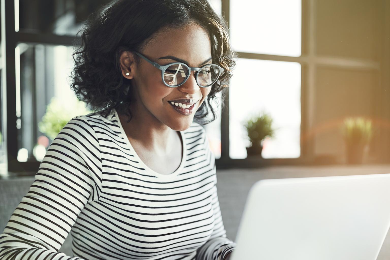 A smiling woman with glasses works on a laptop at a table.