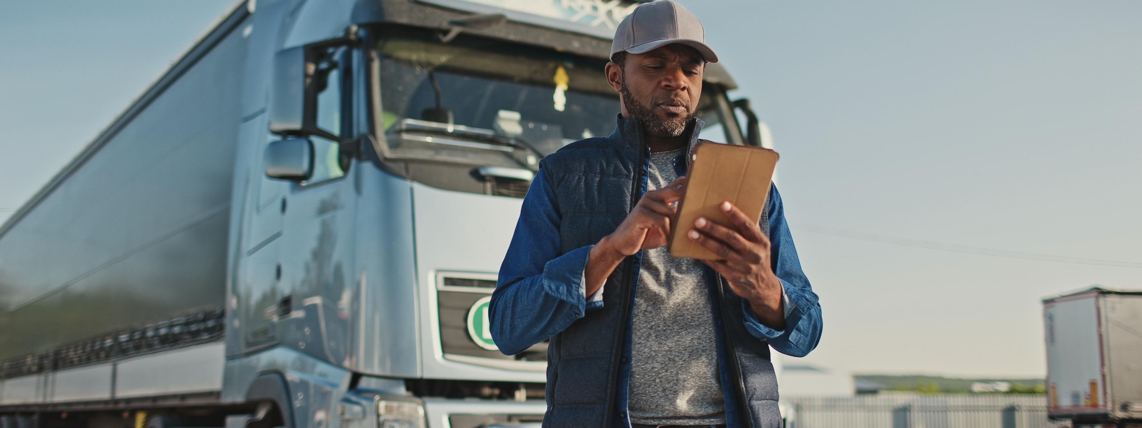 African American professional truck driver setting up navigation for destination. Checking his route on tablet computer and standing by long vehicle. Transportation service.