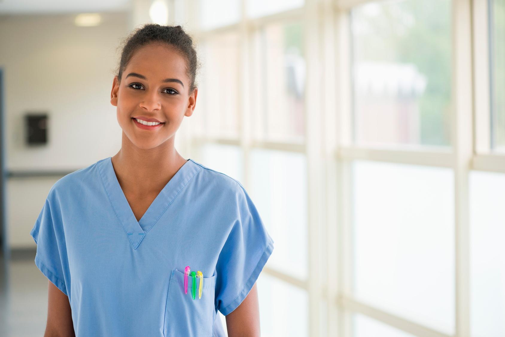 Nurse smiling at camera, standing in sunny hospital room, wearing blue scrubs.