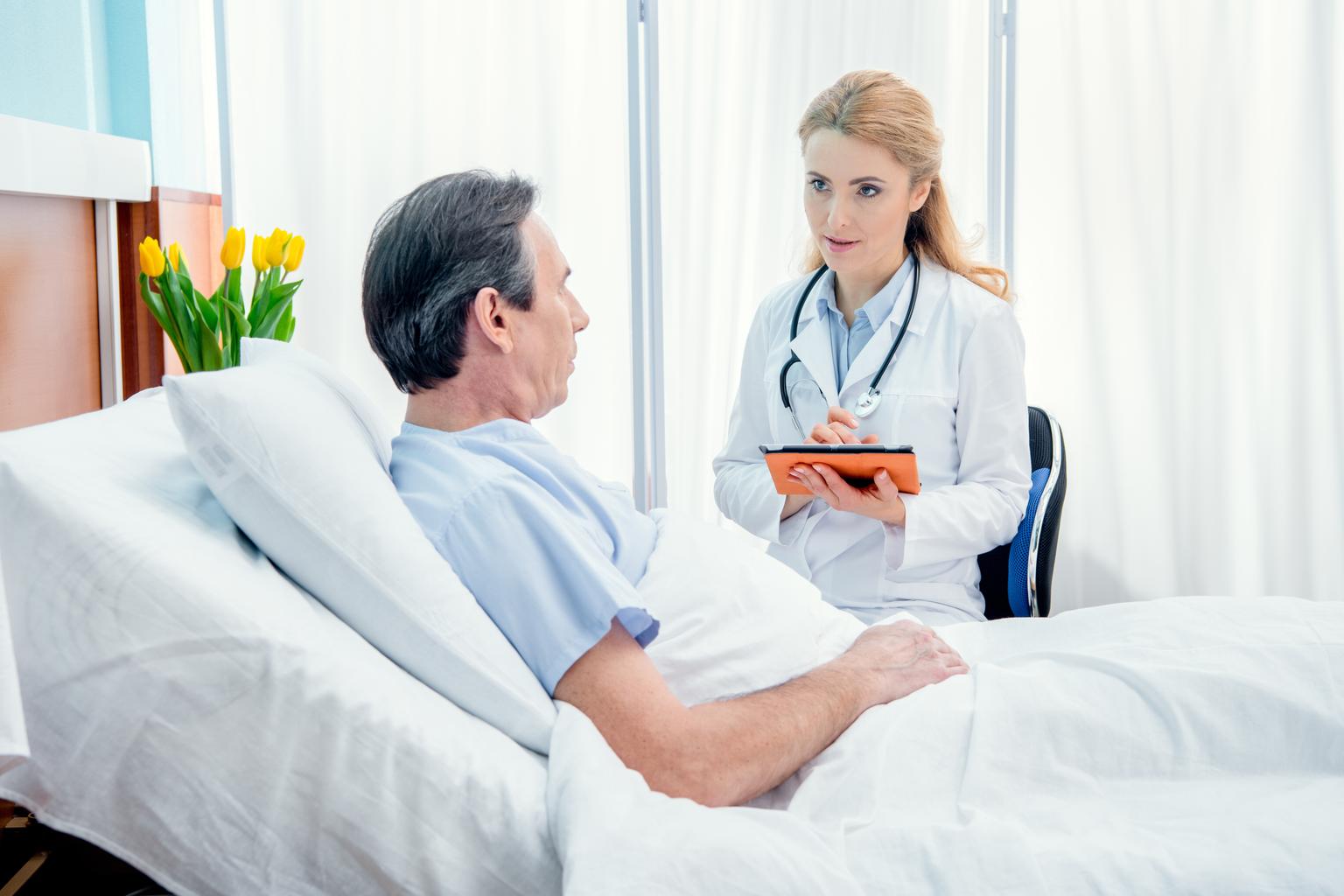 Middle aged male patient lying on hospital bed while getting consultation from female doctor using a tablet