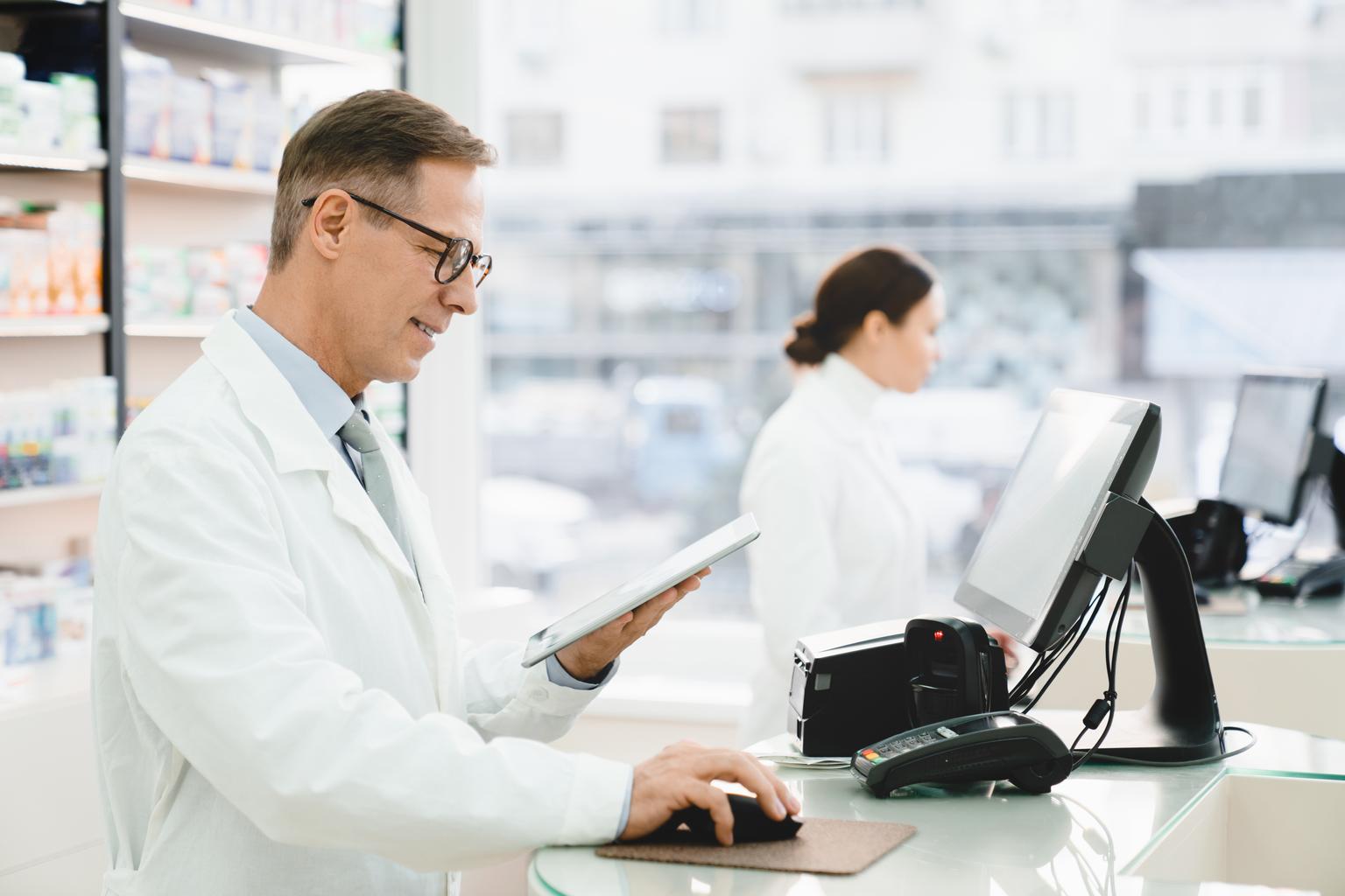 Male and female pharmacists working together at the cash point desk in a pharmacy using computer and tablet