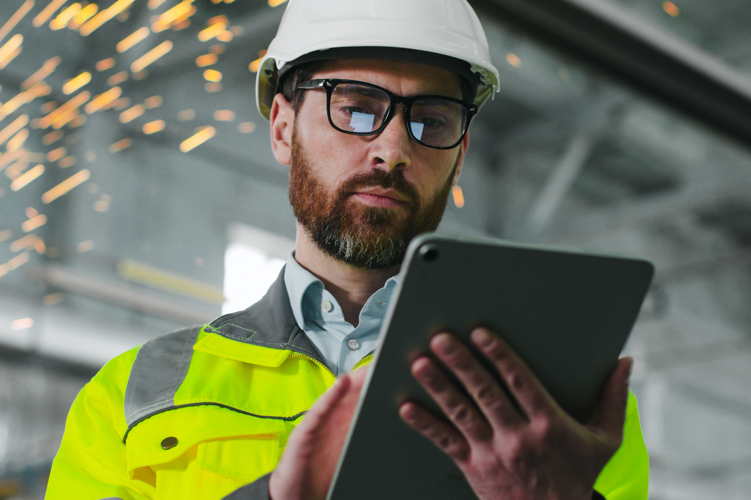 Mature bearded man on construction site. Engineer in uniform and glasses holds tablet background of workflow. An employee notes something in his gadget in workshop. Diversity of proffesions.