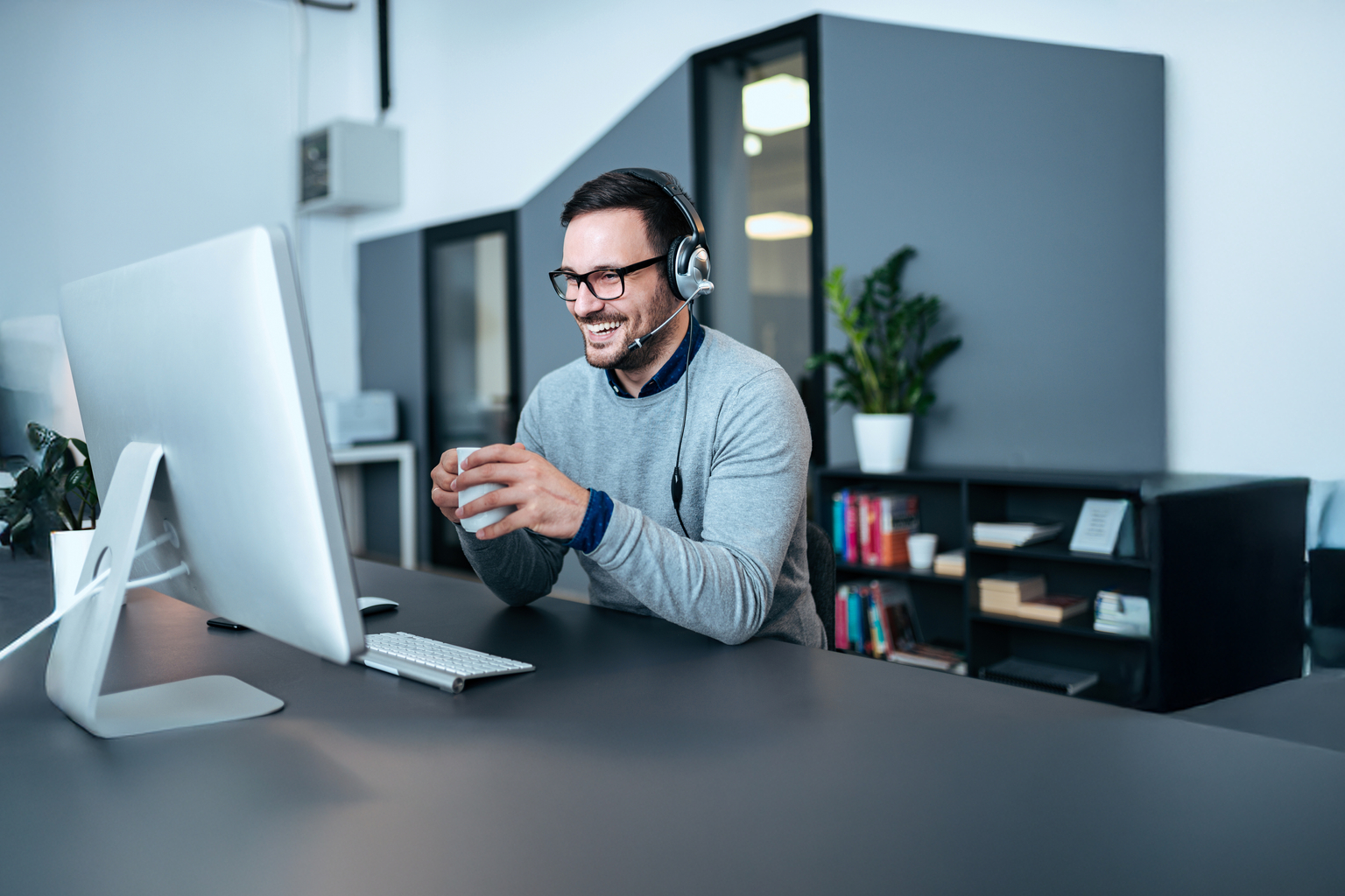 Auf dem Bild ist ein Mann zu sehen, der lächelnd vor einem Computer sitzt mit einer Tasse Kaffee in der Hand.