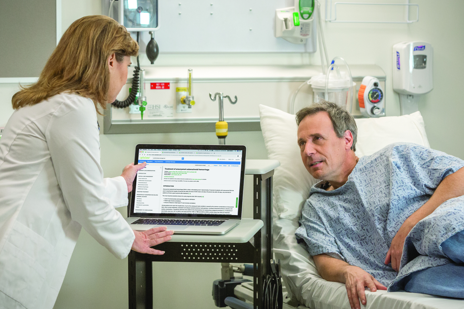 Doctor and patient in hospital room, doctor using UpToDate on laptop