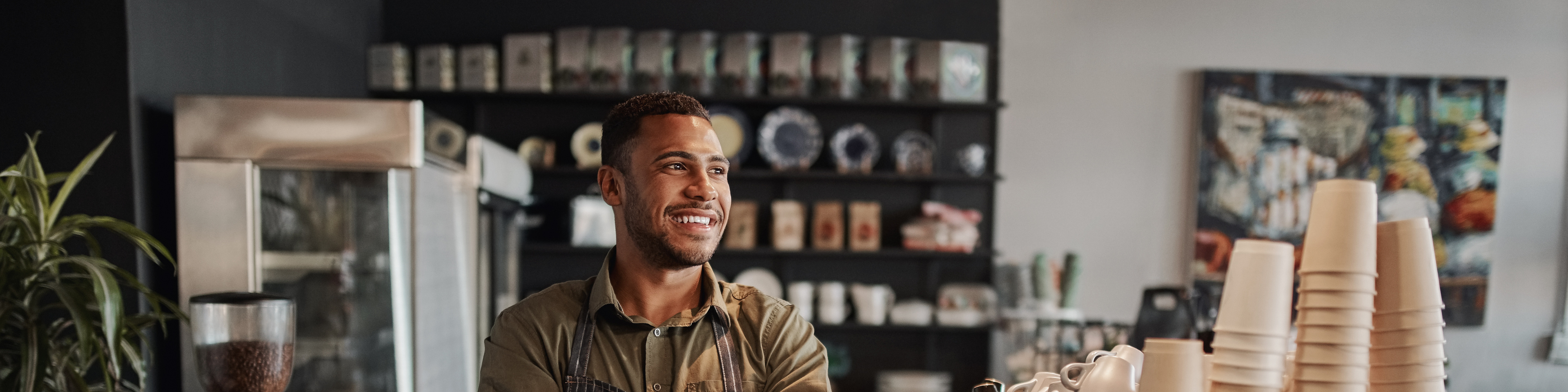 Small coffee shop owner standing behind counter wearing apron with crossed arms