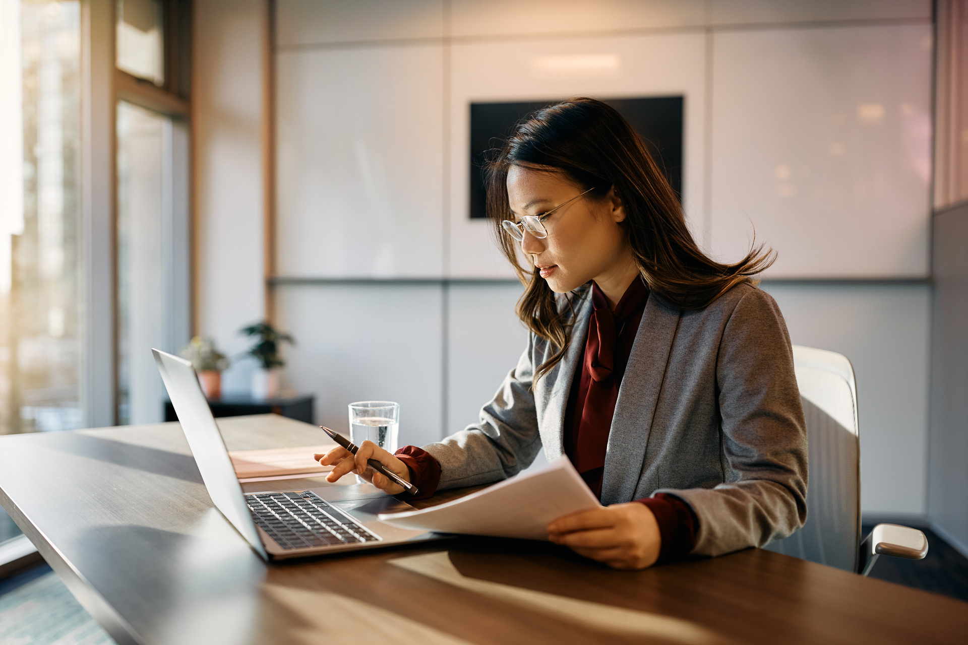 Asian businesswoman using laptop while analyzing reports in office.